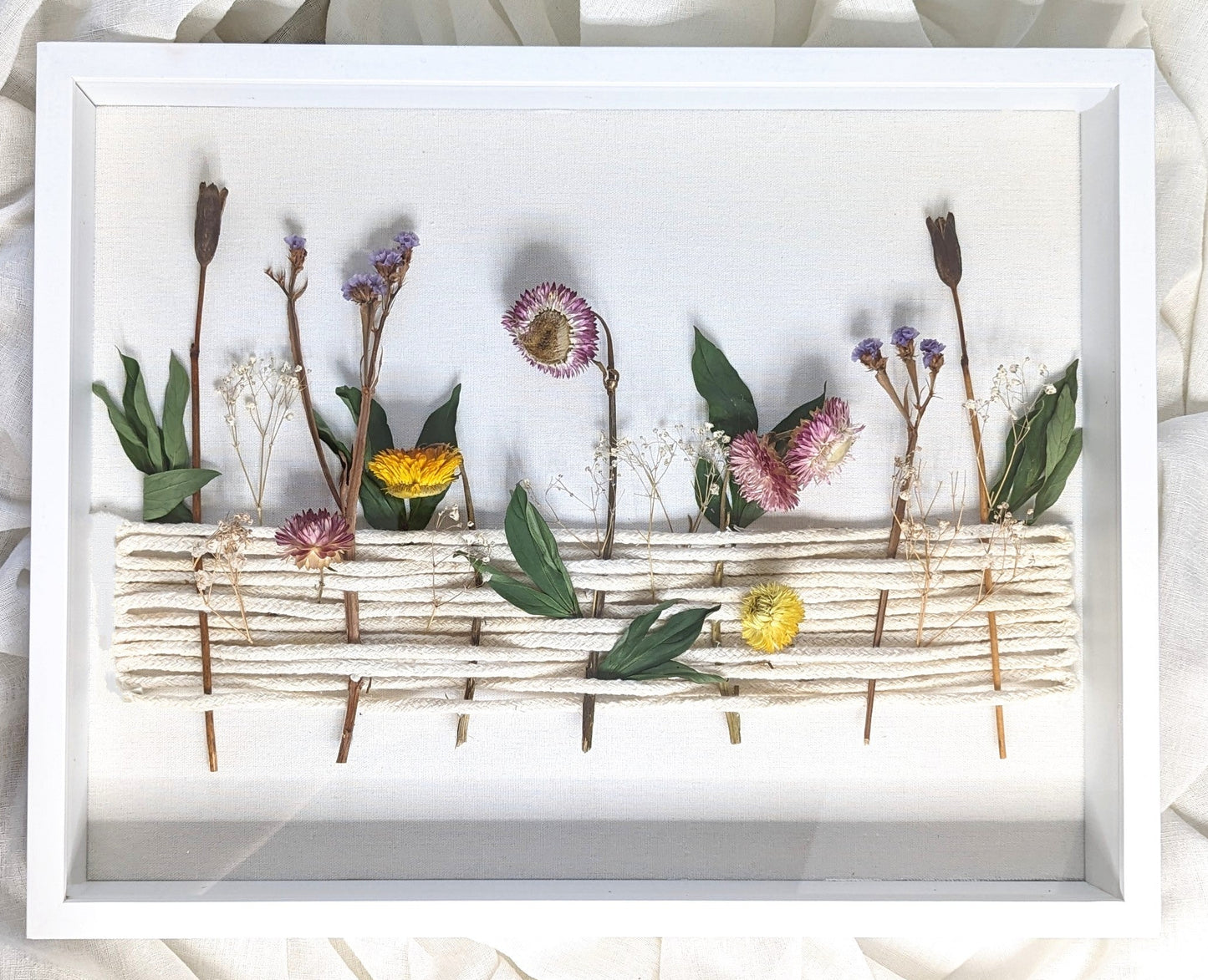 A framed dried flower arrangement featuring various flowers such as daisies and purple accents, set against a white background.