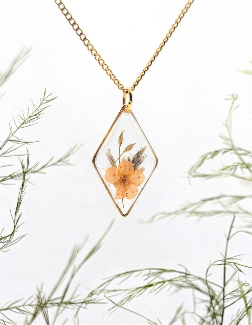 Gold necklace with a diamond-shaped pendant featuring a pressed flower, set against a natural background of green foliage.