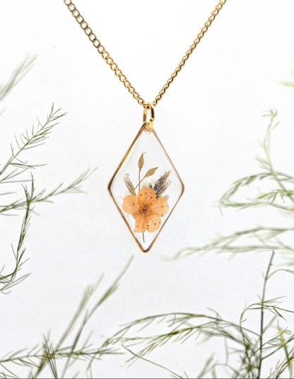 Gold necklace with a diamond-shaped pendant featuring a pressed flower, set against a natural background of green foliage.