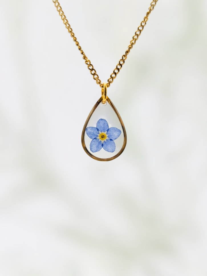 Teardrop-shaped pendant with a blue flower design on a gold chain against a white background