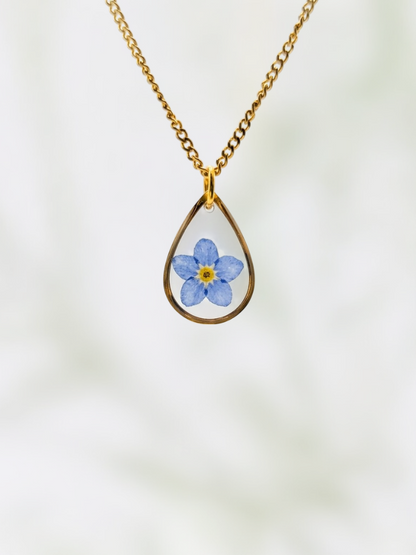 Teardrop-shaped pendant with a blue flower design on a gold chain against a white background
