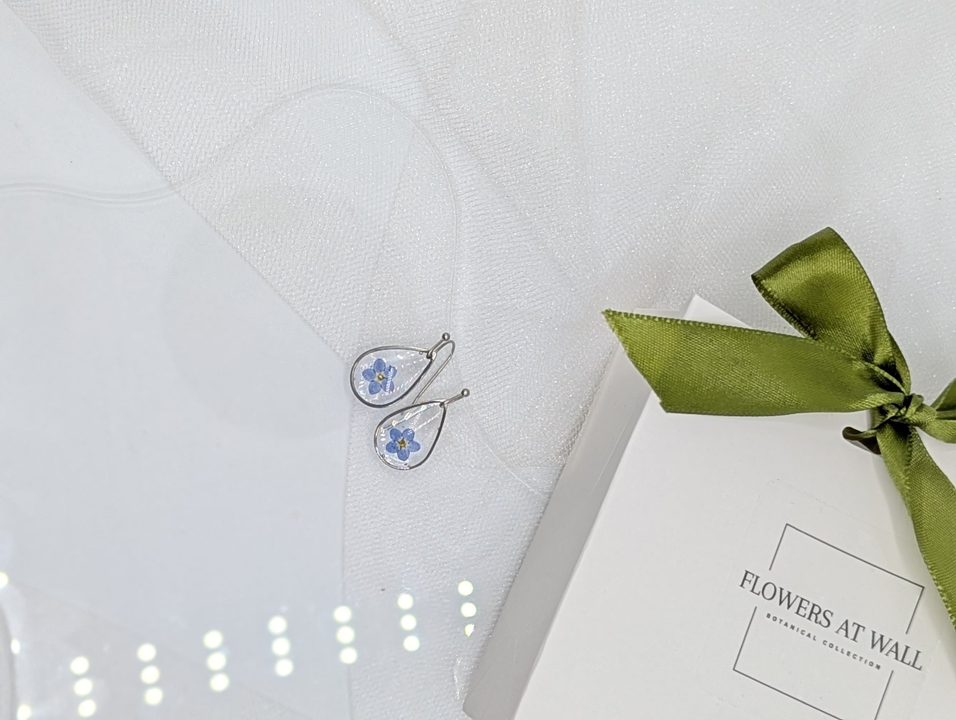 Earrings with floral design on a white box tied with a green ribbon, placed on a textured white surface.
