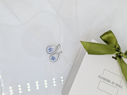 Earrings with floral design on a white box tied with a green ribbon, placed on a textured white surface.