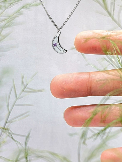 Silver necklace with a crescent moon and purple gemstone held between fingers against a light background with greenery.