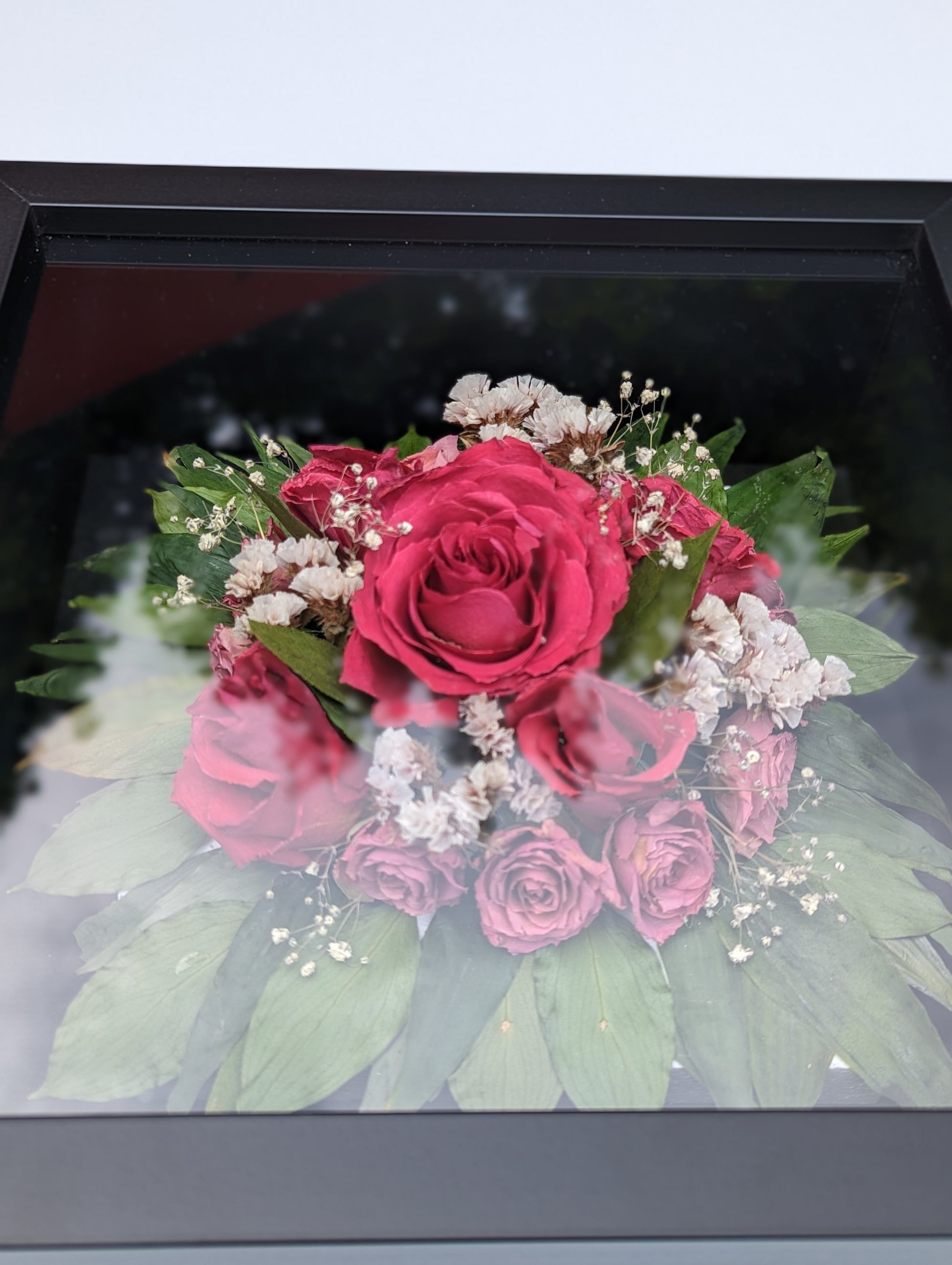 Bouquet of pink roses with green leaves and small white flowers in a black frame.