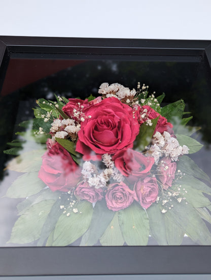 Bouquet of pink roses with green leaves and small white flowers in a black frame.