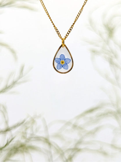 Teardrop-shaped pendant with floral design on a gold chain against a blurred natural background