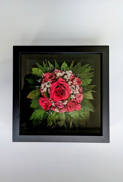 A shadow box bouquet featuring a bouquet of red roses and green leaves, enclosed in a black frame.