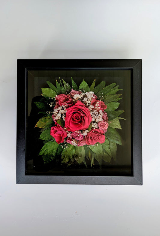 A shadow box bouquet featuring a bouquet of red roses and green leaves, enclosed in a black frame.
